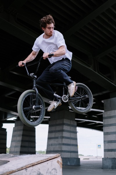 A man in a white shirt doing a stunt airborne with his BMX which has bike pegs on its wheels.