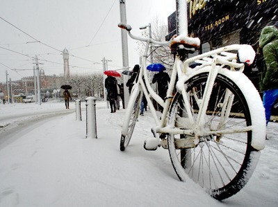 A white bike parked outdoors by a post during winter, on snow.